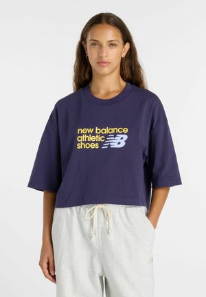Femme aux longs cheveux bruns portant un t-shirt court bleu marine avec un texte New Balance jaune et blanc et un pantalon de jogging gris clair avec cordon.