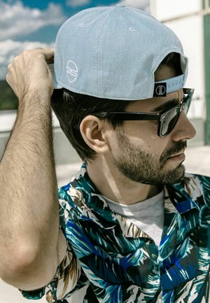 Gorra de béisbol azul claro con logotipo bordado, gafas de sol oscuras y una camisa de pantalón con un patrón tropical en tonos de verde, azul y beige.