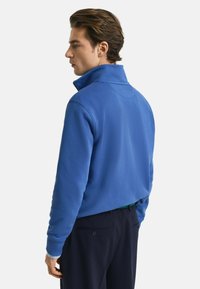 Blaues Sweatshirt mit hohem Kragen und gerippten Bündchen, aus weichem Stoff, von hinten gezeigt. Kombiniert mit dunklen Hosen.