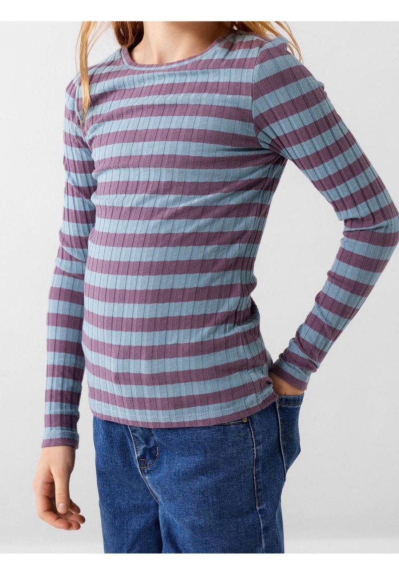Enfant portant un t-shirt à manches longues côtelé avec des rayures horizontales violettes et bleues, et un jean bleu, debout avec une main dans la poche.