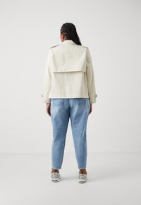 Kremowy płaszcz trencz z klapą z tyłu, zestawiony z jasnoniebieskimi dżinsami i srebrnymi butami sportowymi. Gładka faktura i swobodny fason.