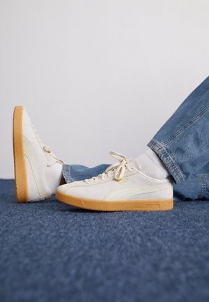 Person iført hvide Puma sneakers med gummisåler, hvide sokker og blå jeans sidder på et blåt tæppe mod en hvid væg.