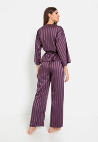 Paarse satijnen pyjamastel met verticale strepen, een ceintuur in de taille en brede mouwen. De top heeft een gebogen zoom en wordt geleverd met bijpassende losse broek.