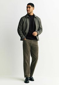Grau strukturierte Jacke über einem schwarzen Pullover, kombiniert mit olivgrünen Cordhosen und schwarzen Loafers. Das Outfit besticht durch ein klares, modernes Design.