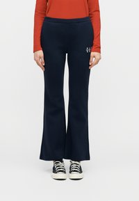 Άτομο που φοράει μπλε σκούρο καμπάνα παντελόνι, μαύρα και λευκά αθλητικά παπούτσια, και πορτοκαλί μακρυμάνικο πουκάμισο, στέκεται μπροστά σε λευκό απλό φόντο.