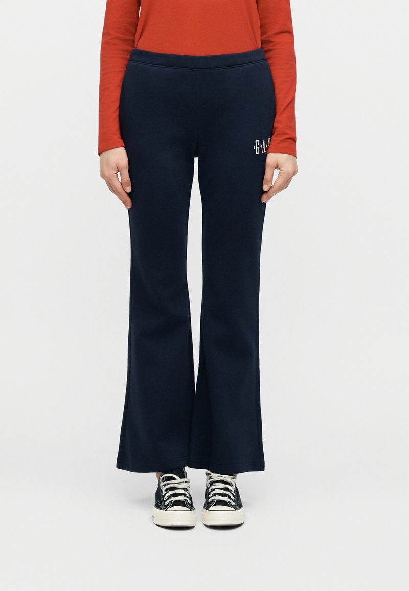 Άτομο που φοράει μπλε σκούρο καμπάνα παντελόνι, μαύρα και λευκά αθλητικά παπούτσια, και πορτοκαλί μακρυμάνικο πουκάμισο, στέκεται μπροστά σε λευκό απλό φόντο.