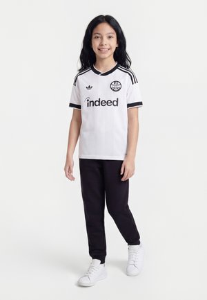 Smilende barn, der står iført en hvid Adidas fodboldtrøje med sorte striber og sorte bukser, hvide sneakers, mod en ensfarvet baggrund.