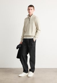 Sweater de malha bege com fecho, apresentando um pequeno logótipo, combinado com calças de ganga escuras e ténis brancos, criando um visual moderno e casual.