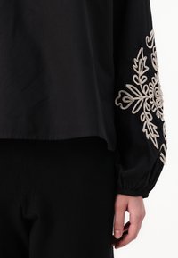 Maglietta nera con vestibilità ampia e maniche lunghe. Presenta un elaborato ricamo floreale beige sulla manica sinistra. Tessuto morbido, design casual.