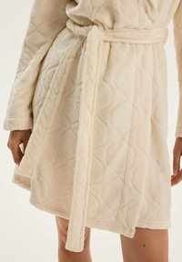 Miękki beżowy fudn rag quilt, związany w talii, noszony przez osobę, której ręce są widoczne, a gołe nogi widoczne poniżej szlafroka.