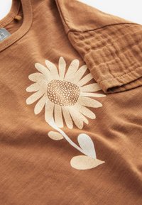 Brūns kokvilnas T-krekls ar teksturētām īsām piedurknēm, kuram ir liela, krēmkrāsas margrietiņa grafika ar sīki izstrādātām ziedlapiņām un lapām baltā krāsā.