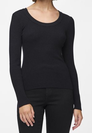 Vrouw draagt een nauwsluitende, geribbelde zwarte trui met lange mouwen en een ronde halslijn, gecombineerd met een zwarte broek tegen een effen achtergrond.