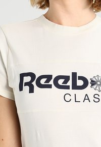 Krátký rukáv, krémové tričko vyrobené z bavlny s kulatým výstřihem, na přední straně s výrazným černým logem "Reebok CLASSIC".