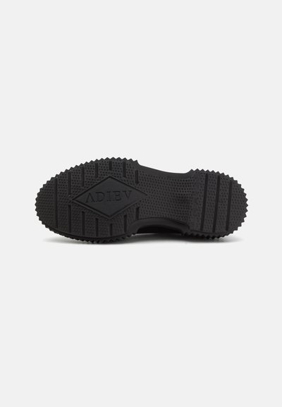 Schwarze Gummisohle mit strukturiertem Grip, die ein diamantförmiges Muster und das eingeprägte Markenlogo zeigt. Abgerundete Kanten und gerippte Muster.