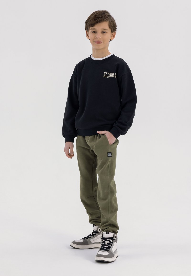 Zwarte sweatshirt met witte ronde hals, groene sweatpants en hoogtop sneakers in grijs en zwart; heeft logo-accenten op de sweatshirt en broek.