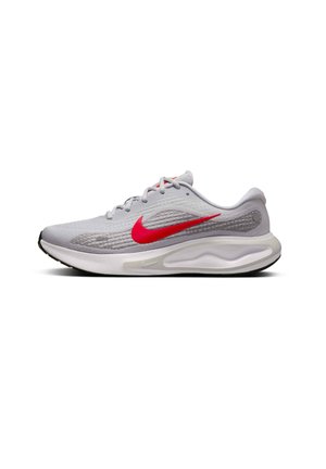 Baskets de sport gris clair avec tige en mesh, swoosh rouge de Nike, et semelle blanche rembourrée. Présente un talon rond et des détails texturés subtils.