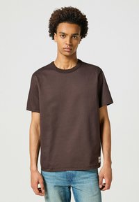 T-shirt in cotone marrone con scollo rotondo e maniche corte. Presenta una linea dritta e un'etichetta piccola sull'orlo. Abbinato a jeans blu chiaro.