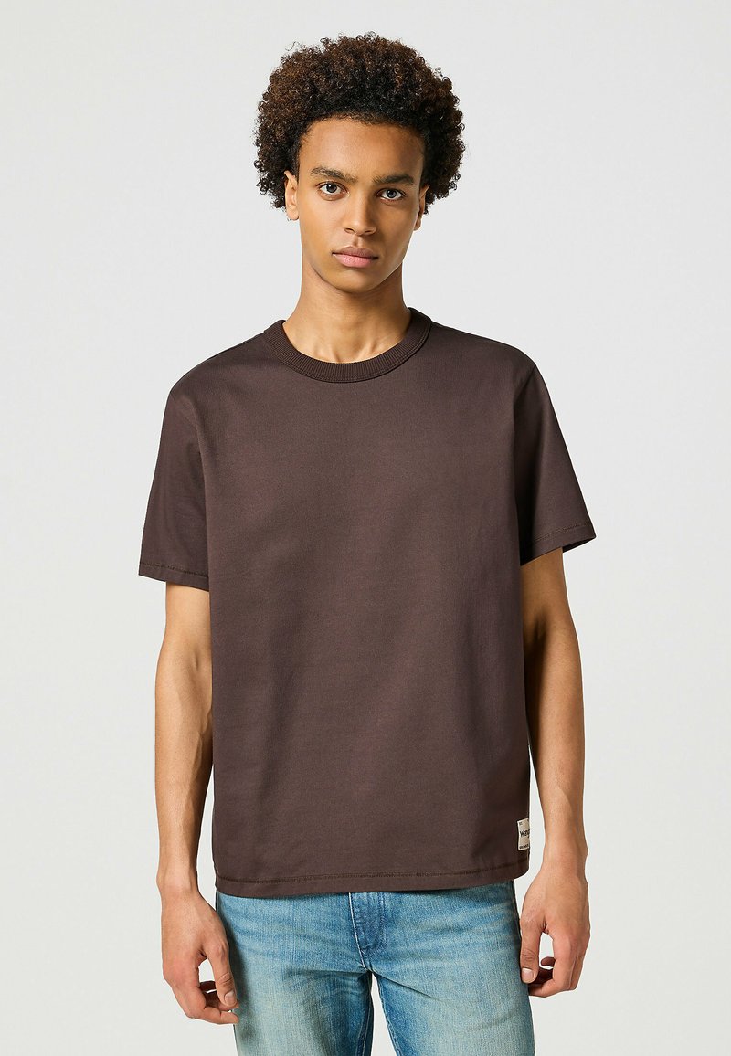 T-shirt in cotone marrone con scollo rotondo e maniche corte. Presenta una linea dritta e un'etichetta piccola sull'orlo. Abbinato a jeans blu chiaro.