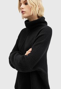 Melna turtleneck džemperis, kas izgatavots no teksturētas auduma, ar brīvu piegriezumu un pazeminātām plecu šuvēm, ar garām piedurknēm un augstu apkakli.