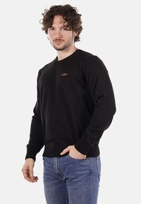 Maglione nero con maniche lunghe, scollo rotondo e polsini a coste. Presenta un piccolo emblema dorato sul petto. Abbinato a jeans blu.
