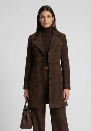 Frau trägt einen dunkelbraunen Wildledermantel über einem Rollkragenpullover, hält eine passende braune Handtasche und steht vor einem schlichten hellen Hintergrund.