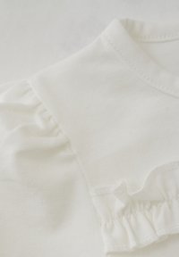 Weißes Baumwolloberteil mit rundem Ausschnitt, kurzen gerafften Ärmeln und einer Rüsche entlang der Schulternaht, mit weicher, glatter Textur.