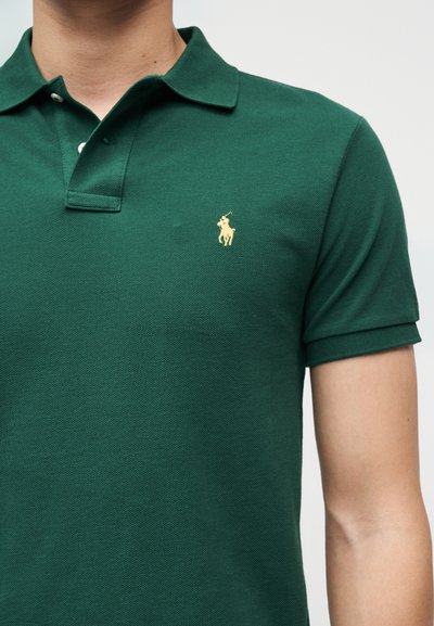 Άνδρας που φοράει σκούρο πράσινο πόλο με κίτρινο κεντημένο λογότυπο παίκτη πόλο στο αριστερό στήθος, με φόντο απλό.