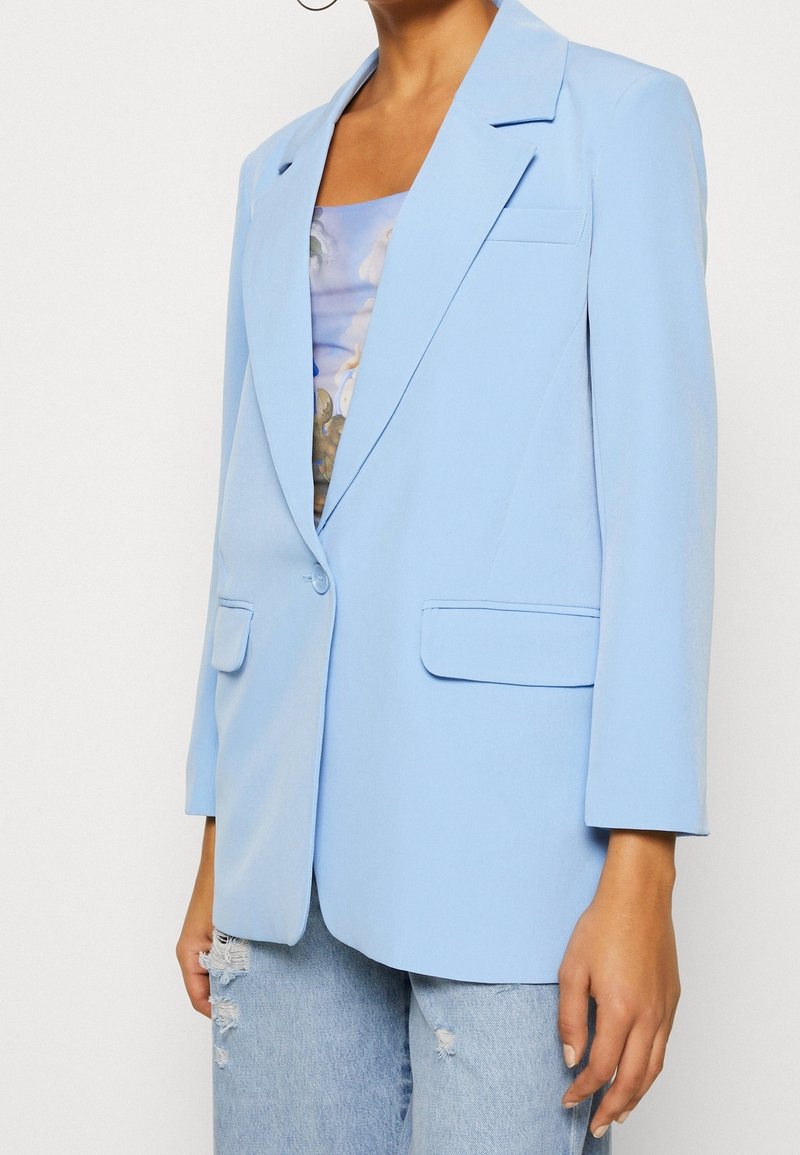 Světlé modré sako s přizpůsobeným střihem, zdůrazněnými rameny a dvěma předními kapsami. Má zapínání na jeden knoflík a hladkou texturu látky.