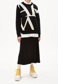 Czarny sweter z dzianiny z kremowym abstrakcyjnym wzorem, noszony na białej koszuli i czarniej spódnicy. Białe sneakersy z żółtymi sznurówkami.