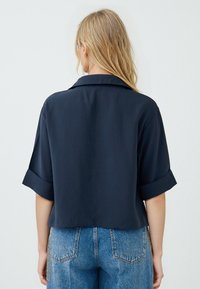 Camicetta cropped blu navy con maniche corte arrotolate e un ampio colletto, realizzata in un tessuto morbido. Abbinata a jeans di denim a vita alta.