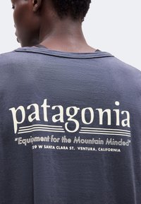 Άτομο που φορεί ένα σκούρο μπλε μπλουζάκι Patagonia με το κείμενο "Equipment for the Mountain Minded" και διεύθυνση Ventura, Καλιφόρνια στην πίσω πλευρά.
