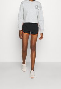 Sudadera gris cortada con logo, combinada con shorts negros con ribete naranja. Lleva puestas unas zapatillas deportivas de color claro.