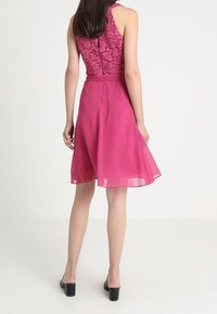 En knälång klänning med en åtsittande spetsöverdel och en flödande, genomskinlig rosa kjol, med en dragkedja i ryggen och en öppen ryggdesign. Enkla svarta klackar.