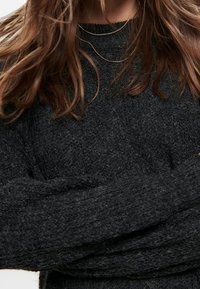 Maglione in maglia grigio scuro con un fondo testurizzato, caratterizzato da maniche lunghe e colletto a girocollo, indossato sopra sottili collane d'oro.