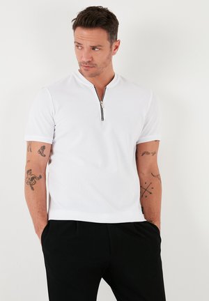 Camisa polo blanca de manga corta con cuello con cremallera, tejido texturizado y diseño ajustado, combinada con pantalones negros.