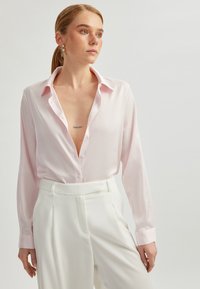 Light pink blouse in smooth fabric, features a deep V-neck, button closure, and long sleeves; paired with high-waisted white trousers.