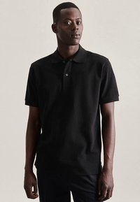 Zwart poloshirt gemaakt van katoen, voorzien van een kraag, korte mouwen en een placket met drie knopen, gedragen door een model dat staat.