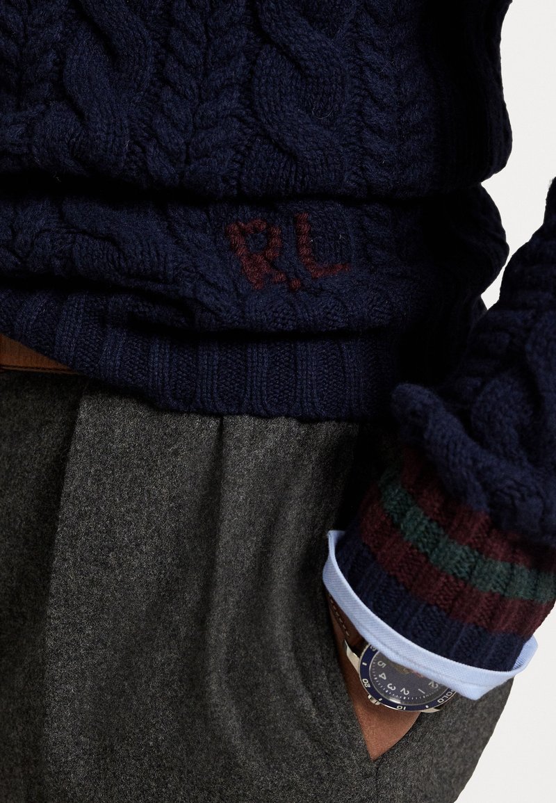 Maglione navy a trecce con polsini a coste in rosso scuro e accenti verdi. Iniziali ricamate "R.L." visibili sull'orlo, abbinato a tessuto grigio.