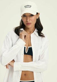 Biała czapka baseballowa z napisem "PARIS TENNIS CLUB" na czarno, w połączeniu z rozpiętą białą koszulą na czarnym bikini z białym wykończeniem.