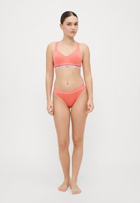 Coral pink bikini set with a V-neck bralette and matching high-waisted bottoms, featuring a logo band at the waist and smooth texture.