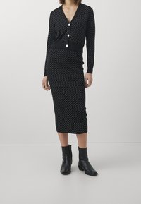 Vrouw in een zwart polka dot vest met lange mouwen en bijpassende midi rok, met zwarte enkellaarsjes, staande tegen een effen achtergrond.