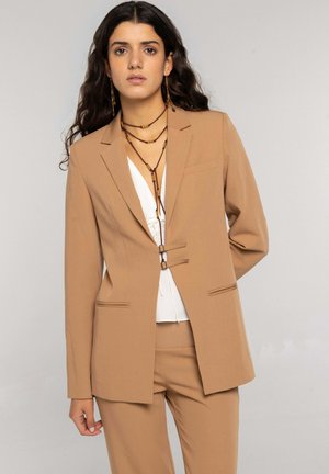 Mujer con cabello largo, rizado y oscuro, vestida con un traje color camello, blusa blanca y collares marrones de cuentas en capas, de pie frente a un fondo neutro.