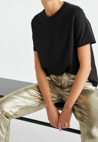 Sort bomulds t-shirt med korte ærmer, kombineret med metallisk guld bukser, der har en blank tekstur og synlige syninger.