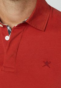 Primo piano di una persona che indossa una maglietta polo rossa con un colletto interno a scacchi in bianco e nero e un piccolo logo ricamato sul petto.