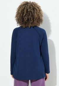 Top navy a maniche lunghe con una vestibilità comoda, maniche raglan e una texture morbida. Dettagli delle cuciture visibili e orlo curvo. Abbinato a pantaloni viola.