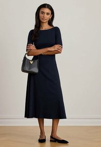 Donna in un vestito midi blu navy con maniche a tre quarti, che tiene una borsa nera, indossa delle slippers nere con cinturino, in piedi su un pavimento di legno.