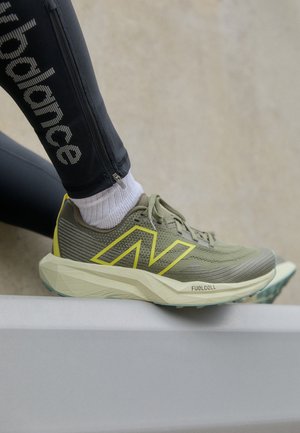 Atletsko sko med gule accenter og "FuelCell"-sål, båret med hvide sokker og sorte New Balance leggings, fod hvilende på en kant.