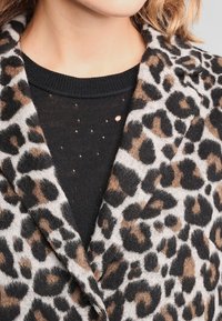 Mujer vestida con un top negro de punto con pequeños agujeros y un abrigo de pelaje con estampado de leopardo encima, enfocándose en el área del escote y el pecho.