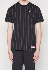 Černé tričko s krátkým rukávem a bílými svislými pruhy, bílé logo Jumpman na levé hrudi a malá červená obdélníková nášivka blízko lemu.