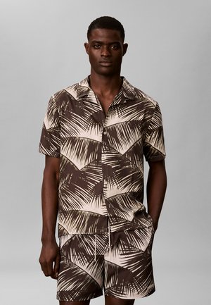 Mand iført matchende brun og beige skjorte og shorts med palmebladtryk, stående mod en ensfarvet grå baggrund.